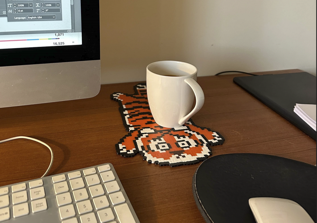 A cute, stylised tiger-shaped coaster sits on a desk with a mug on top of it. A computer screen, keyboard, mouse and notepad surround the coaster.