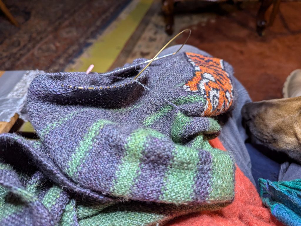 A green and grey striped knitted item is resting on a lap, as seen from above. Circular knitting needles are holding stitches around part of the garment, and a cross stiched tiger is partially visible to one side. A tan dog's nose is resting on the seat next to the person holding the knitting.