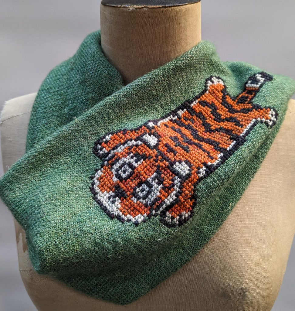 A dressmakers dummy has a green knitted cowl draped around its neck. The cowl has a cute, stylised tiger cross stitched along the length of the cowl.