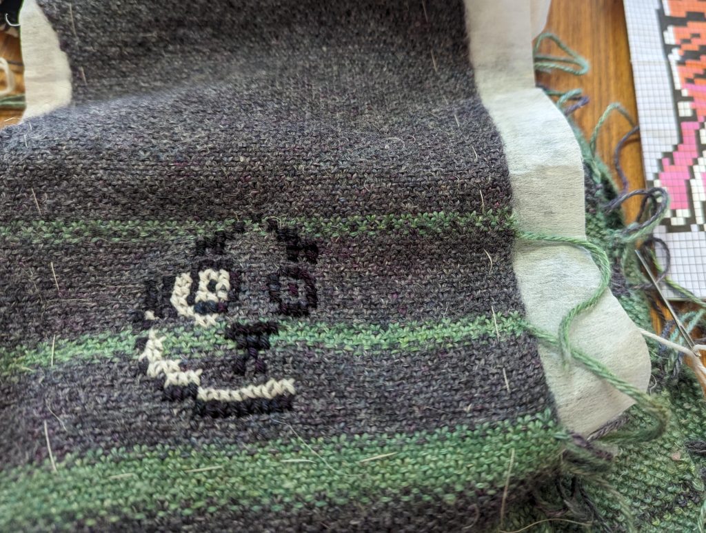 A green and grey striped, knitted fabric has a partial face - with two eyes, a nose and part of a cheek - emerging in cross stitch. White, tear-away stabiliser that is hand basted to the back of the knitting is poking out at the sides. A needle and thread, and part of a chart showing the orange, black and white of a tiger is visible to the right of the image.