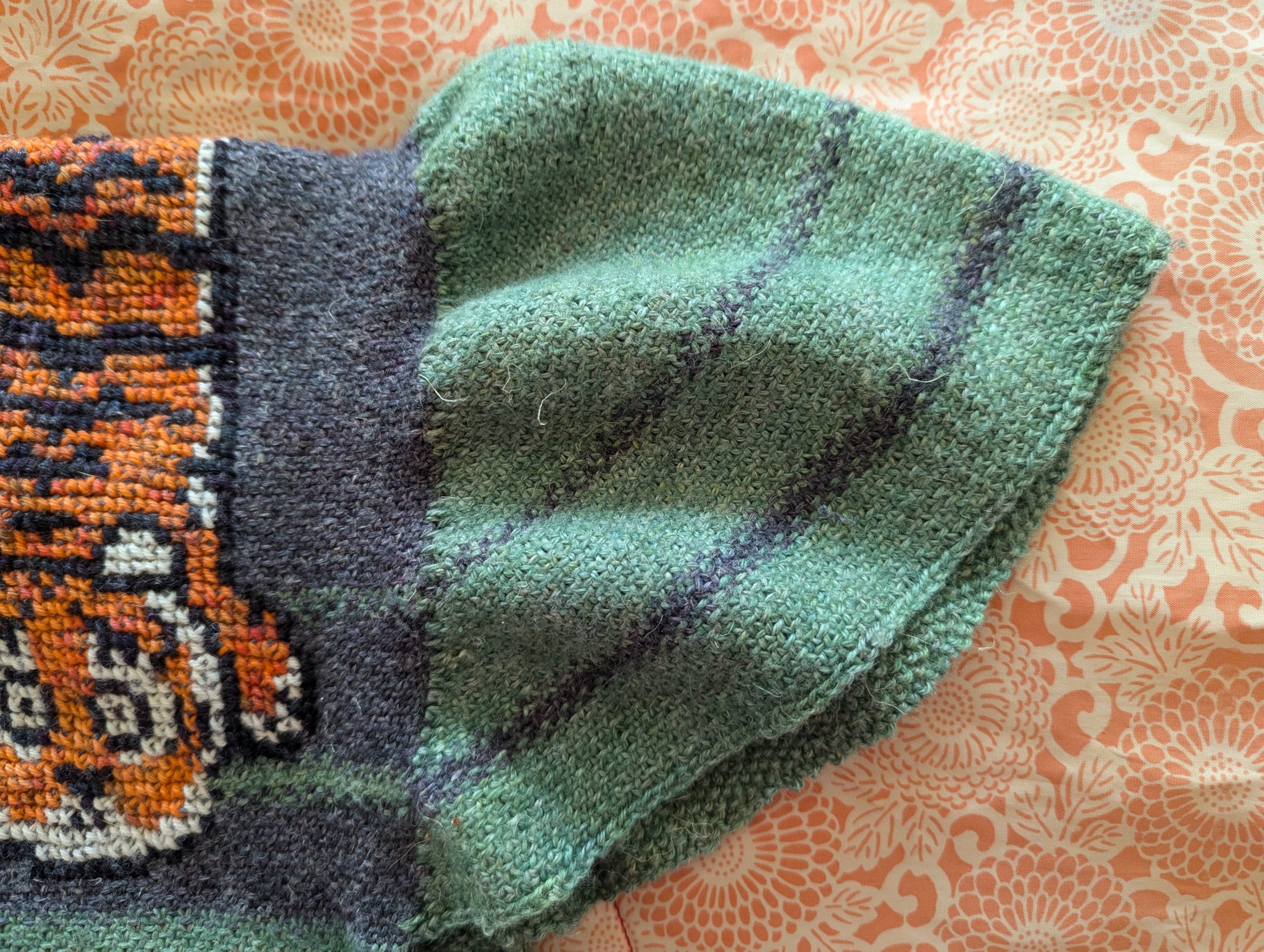 The puffed sleeve and shoulder of a grey and green knitted top sits on a pale orange and cream patterned fabric. The shoulder has a tiger motif, and the sleeve has stripes.